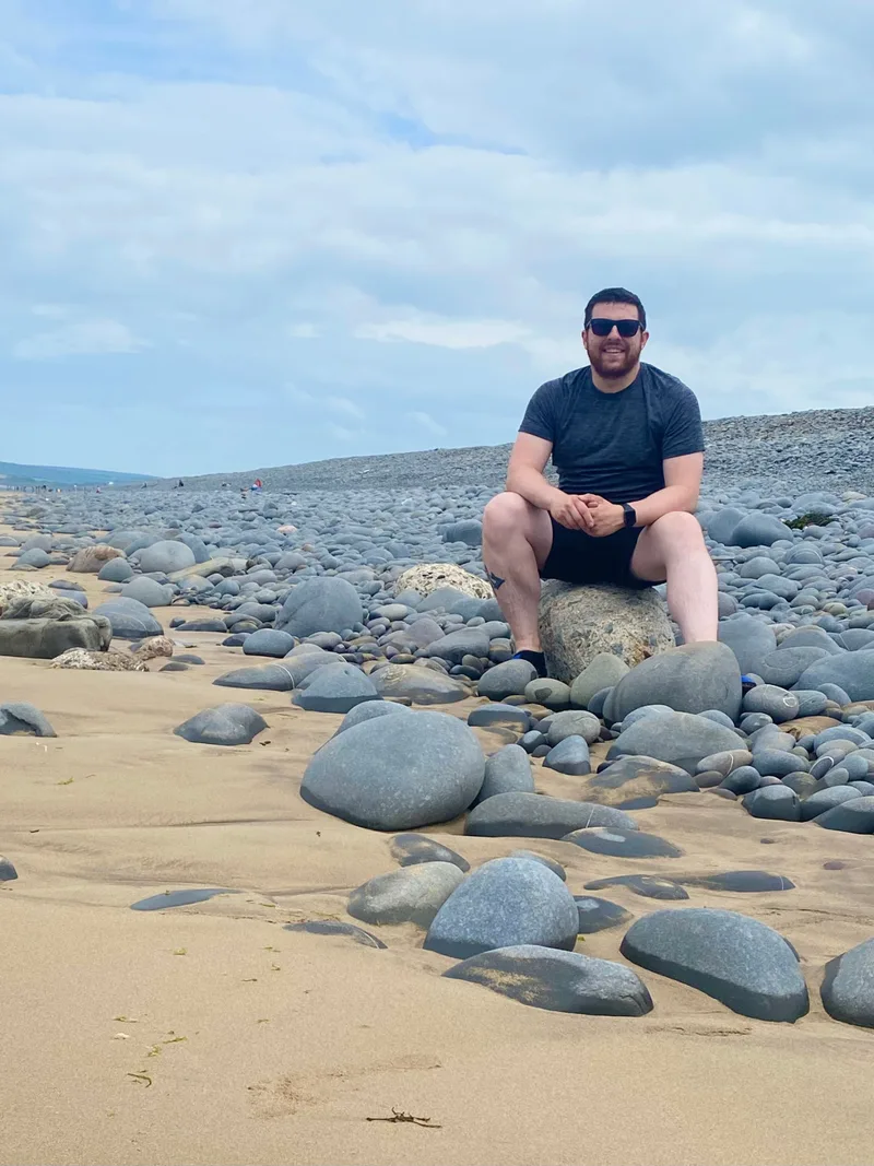 Dale Harris smiling, sitting on a rock on a pebble beach